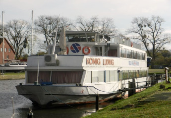 Ein Foto eines Hausbootes namens KÖNIG LUDWIG, das an einem ruhigen Kanal mit Bäumen und Wiese liegt.