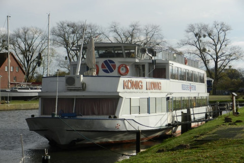 A photo of a houseboat named KÖNIG LUDWIG moored along a calm canal, surrounded by trees and grass.
