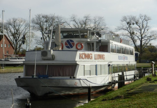 A photo of a houseboat named KÖNIG LUDWIG moored along a calm canal, surrounded by trees and grass.