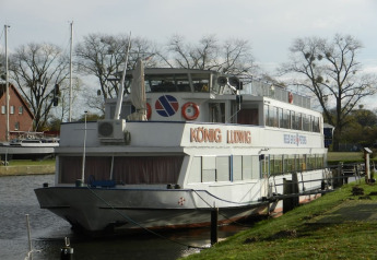 Foto de una casa flotante llamada KÖNIG LUDWIG atracada en un canal tranquilo, rodeada de árboles y césped.