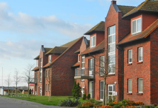 Modern brick apartment buildings with large windows and balconies, lined with trees and flowerbeds outside.