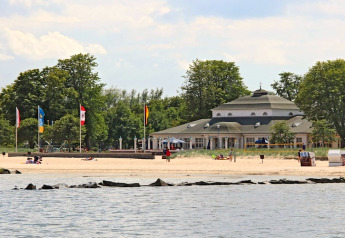 Zicht op een zandstrand met enkele mensen, een opvallend gebouw en bomen op de achtergrond aan het water.