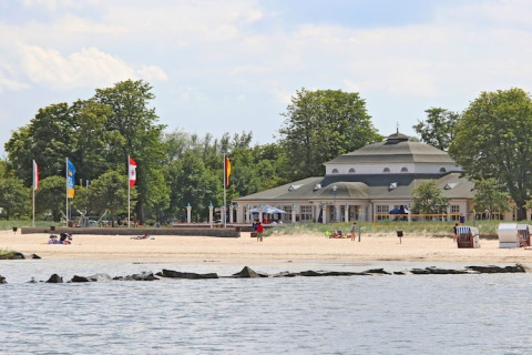 View of a sandy beach with a few people, a distinctive building, and trees in the background by the water.
