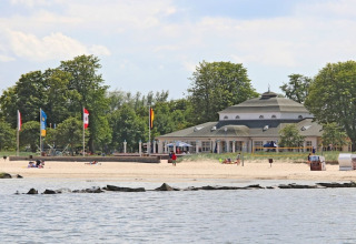 View of a sandy beach with a few people, a distinctive building, and trees in the background by the water.