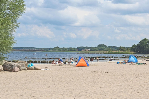 Beach scene with people, small tents, sandy shore, and a partly cloudy sky by the sea.