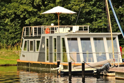 Modern houseboat with large windows and rooftop deck with parasol, docked at Houseboat Xenia in Germany.