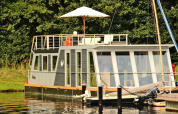 Moderne woonboot met grote raampartijen en dakterras met parasol, aangemeerd bij Houseboat Xenia in Duitsland.