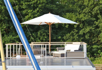 Dakterras van woonboot 'Xenia' in Duitsland, met loungeset, parasol en zicht op groene bomen.