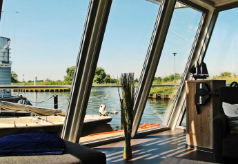 Blick aus dem Hausboot «Xenia» in Deutschland mit großen Fenstern, Sofa und Blick auf Hafen und Natur.