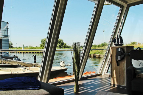 View from Houseboat ‘Xenia’ in Germany with large windows, a sofa, and a scenic river and harbor outside.