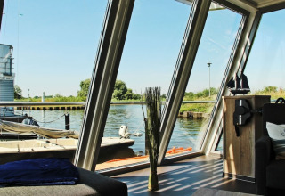 View from Houseboat ‘Xenia’ in Germany with large windows, a sofa, and a scenic river and harbor outside.
