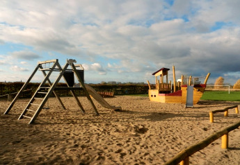 Speelplein op zand met schommels, een glijbaan en een speelboot onder een half bewolkte hemel.