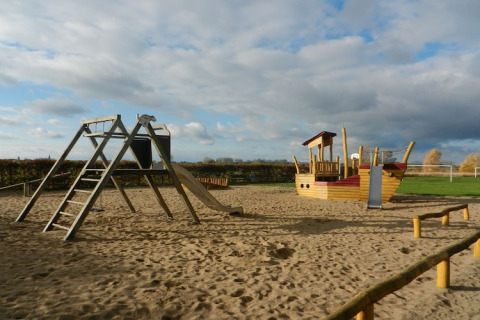 Playground on sand with swings, a slide, and a wooden ship playhouse under a partly cloudy sky.