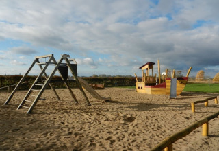 Playground on sand with swings, a slide, and a wooden ship playhouse under a partly cloudy sky.