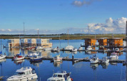Casa flotante y barcos en un puerto tranquilo con cielo azul y campos verdes al fondo en el paisaje.