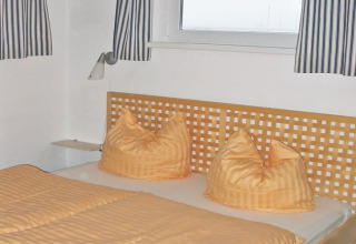 Slaapkamer op een woonboot met oranje beddengoed, gestreepte gordijnen en houten latten hoofdbord.