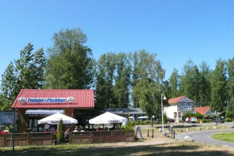 Udendørs café med parasoller og et rødt tag, omgivet af træer under en blå himmel på en solrig dag.