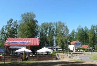 Buitencafé met rood dak en parasols, omringd door bomen en een blauwe lucht op een zonnige dag.