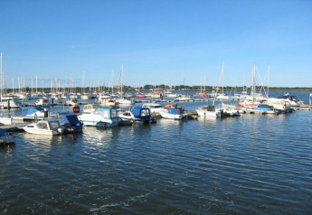 Både og husbåde ligger fortøjet ved en marina på en solrig dag med klart blåt vand og himmel.