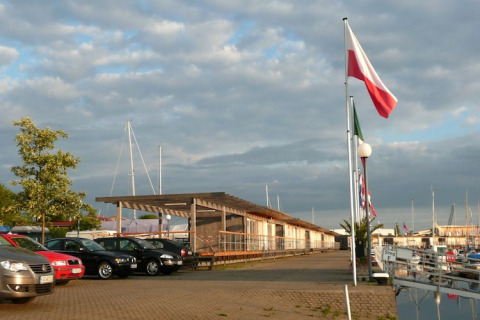Boats and parked cars near houseboats at a marina, flags waving under a partly cloudy sky at sunset.