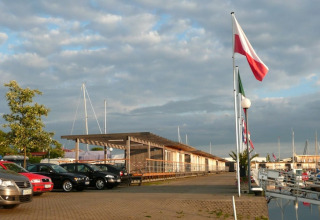 Boten en geparkeerde auto’s bij woonboten aan de haven, vlaggen wappert onder deels bewolkte lucht.