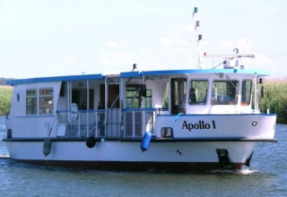 Et hvidt husbåd med navnet Apollo I sejler på en flod omgivet af grøn vegetation under en klar himmel.