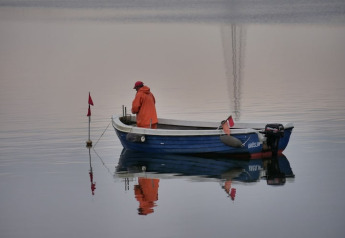 Persoon in oranje regenjas op een kleine blauwe motorboot op rustig water, met reflecties.