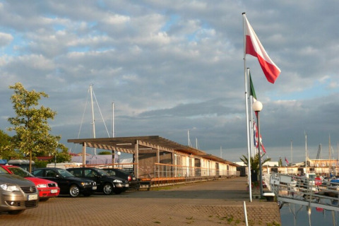 Afbeelding van een woonboot-haven met auto's, vlaggen, aanlegsteiger en boten onder bewolkte hemel.