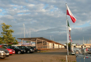 Foto van een woonboot-haven met geparkeerde auto's, vlaggen en boten onder een bewolkte lucht.