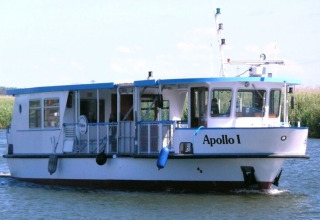 A photo of a houseboat named Apollo I cruising on the water, with greenery in the background.
