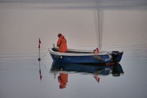 A fisherman in orange rain gear stands on a small blue boat on calm water, surrounded by reflections.