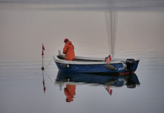 Un pêcheur en ciré orange se tient sur une petite barque bleue sur une eau calme, entouré de reflets.