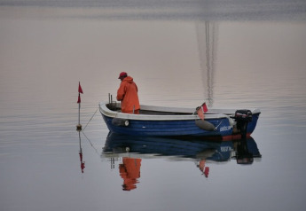 Un pescador con ropa impermeable naranja está de pie en un pequeño bote azul sobre aguas tranquilas.