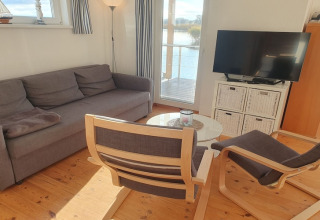 Stue på en husbåd med grå sofa, to stole, rundt bord, TV og vindue med udsigt til vandet.