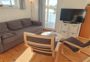 Soggiorno su una casa galleggiante con divano grigio, due poltrone, tavolo in vetro e vista lago.