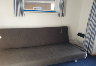 Graues Schlafsofa unter einem Fenster mit blau-weiß gestreiften Vorhängen auf einem Hausboot, blauer Teppich.