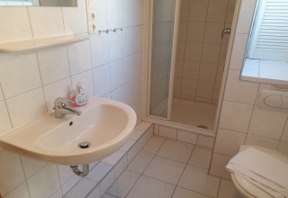 Badeværelse på en husbåd med vask, bruser, toilet og spejl, alt i lyse fliser og moderne stil.