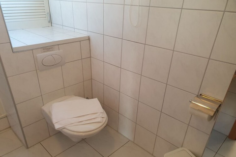 A houseboat bathroom with white tiles, a wall-mounted toilet, a folded towel, and a toilet paper roll.