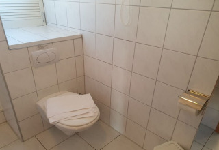 A houseboat bathroom with white tiles, a wall-mounted toilet, a folded towel, and a toilet paper roll.