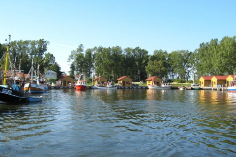 Photo d’un port paisible avec des péniches et des bateaux de pêche devant des arbres et un ciel bleu.