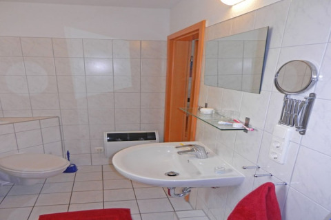 Badkamer op een woonboot met witte tegels, hangtoilet, spiegel, wastafel en een rode handdoek.