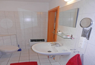Badkamer op een woonboot met witte tegels, hangtoilet, spiegel, wastafel en een rood handdoekje.