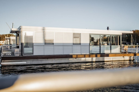 Moderne woonboot met grote ramen en terras, aangemeerd in een haven in Duitsland tijdens daglicht.