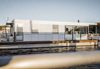Moderne woonboot met grote ramen en terras, aangemeerd in een haven in Duitsland tijdens daglicht.