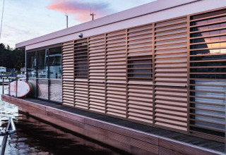 Moderne woonboot met grote lamellen en glazen panelen, aangemeerd in een Duitse haven bij zonsondergang.