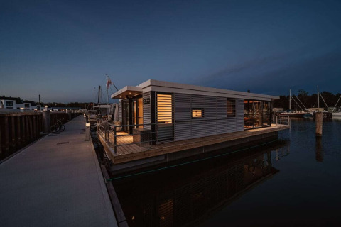 Moderne woonboot aangemeerd in een Duitse jachthaven, 's avonds verlicht en aan de steiger verbonden.