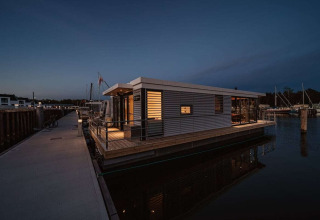 Moderne woonboot aangemeerd in een Duitse jachthaven, 's avonds verlicht en aan de steiger verbonden.