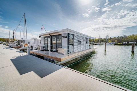 Moderne woonboot genaamd «475077» aangemeerd in een Duitse jachthaven, omringd door water en blauwe lucht.
