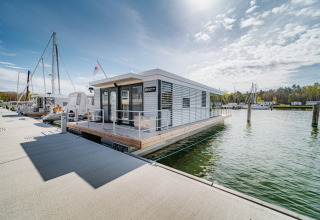 Moderne woonboot genaamd «475077» aangemeerd in een Duitse jachthaven, omringd door water en blauwe lucht.