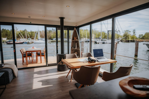 Modern houseboat interior in Peenemünde with workspace, terrace seating, and marina view outside windows.